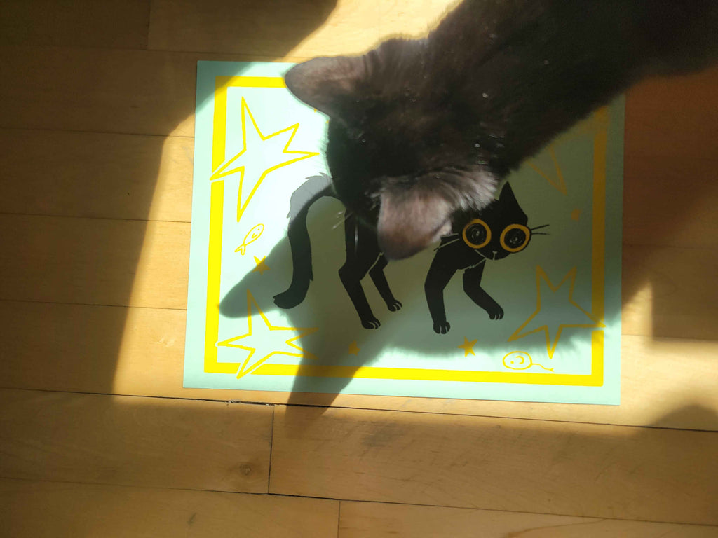 Black cat interacting with a screenprinted illustration of a black cat with yellow eyes.